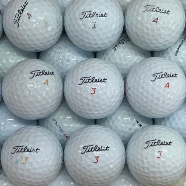 A close-up pile of white Titleist golf balls, with several visible in the center showing the black Titleist logo and red numbered markings.