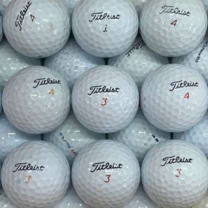 A close-up pile of white Titleist golf balls, with several visible in the center showing the black Titleist logo and red numbered markings.